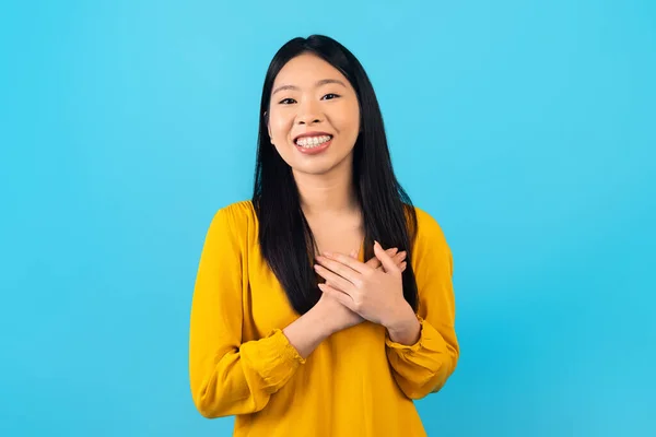 积极感恩的年轻韩国女人 身穿黄色衬衫 双手捂住胸膛 对着摄像机微笑 表达爱意 与蓝色背景隔离 — 图库照片