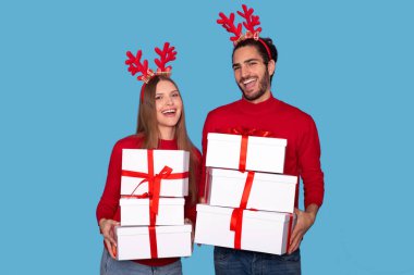 Ren geyiği boynuzları taşıyan neşeli genç çift, Noel hediyeleri taşıyan mutlu adam ve kadın, kameraya gülümseyen, mavi arka planda izole edilmiş pozlar veren,