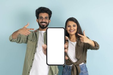 Harika bir mobil uygulama. Heyecanlı Hintli çift, dev akıllı telefonu sunuyor. Uygulamayı veya web sitesini tanıtmak, reklam ürünü veya servisi, mavi arka plan, stüdyo çekimi.