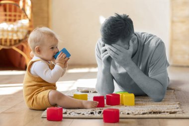 Ebeveyn tükenmesi. Depresyondaki baba, bebek bebekle yan yana otururken üzgün genç adam, küçük çocuğa bakmaktan, babalık izni sırasında ruhsal kriz yaşamaktan, kolaj yapmaktan sıkılmış.