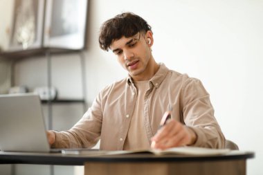 E-Öğrenme Teklifi. Arap öğrenci laptopta notlar yazıyor evde kablosuz kulaklık takıyor masada oturuyor ve Webinar 'dan bilgi alıyor. Teknoloji ve Öğrencilik