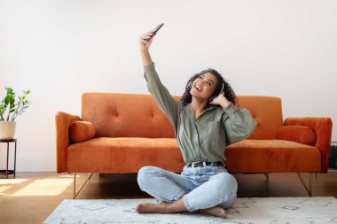 Mutlu genç siyahi bayan, kablosuz kulaklıkla selfie çekiyor, evde online müzik dinliyor, modern oturma odasının içinde yerde oturuyor. Sosyal Medya, Blogger Yaşam Biçimi