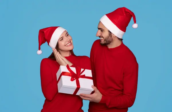 Gift For You. Young Man In Santa Hat Making Christmas Surprise To His Girlfriend, Giving Present Box To Excited Beautiful Woman, Loving Couple Standing Isolated On Blue Background, Copy Space