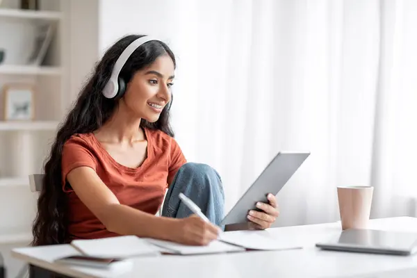 Student Young Indian Woman Have Online Learning Session At Home. Cheerful millennial lady sitting at desk with digital tablet, using headset, taking notes, attend online course, webinar, copy space