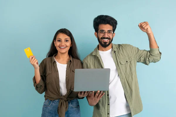 Positive happy millennial indian man and woman couple spouses posing with laptop and bank credit card isolated on blue plain background, gesturing and smiling. Cashback, online shopping and banking