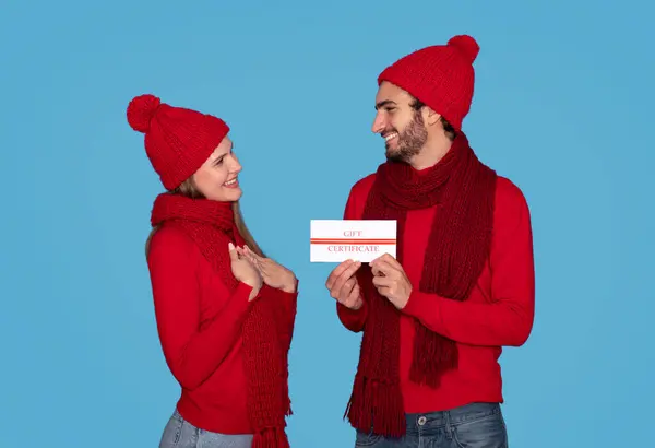 Loving Man Presenting Gift Certificate To His Happy Girlfriend, Romantic Young Couple In Knitted Hats Celebrating Winter Holidays Together, Standing Isolated On Blue Studio Background, Free Space