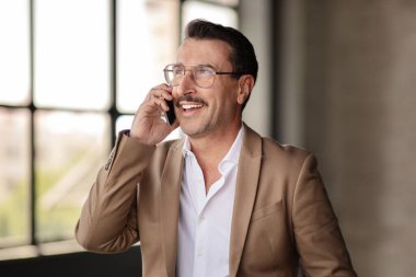 İş İletişimi. Başarılı, olgun bir iş adamı iş yerinde cep telefonuyla sohbet ediyor. Gülümseyen girişimci portresi cep telefonuyla arıyor ve konuşuyor.