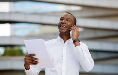 Gülümseyen siyahi iş adamı cep telefonuyla konuşuyor ve dışarıda belgeler okuyor, beyaz gömlekli mutlu Afrikalı Amerikalı erkek girişimci iş ortağıyla telefonda konuşuyor, Boşluğu Kopyala