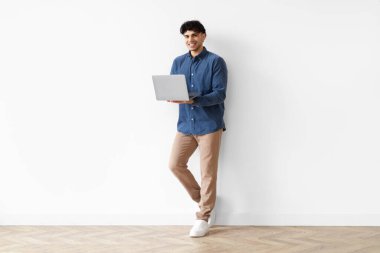 Orta Doğulu neşeli adamın dizüstü bilgisayarı tutması ve bilgisayarla çalışması beyaz duvara yaslanarak kameraya gülümsemesi. Genç Freelancer Reklamcılık Web Sitesi