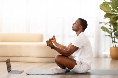 Yakışıklı genç zenci adam evde meditasyon yapıyor, dizüstü bilgisayar kullanıyor, spor minderinde lotus pozu veriyor, internette video izliyor, kopyalama alanı, yan görüş. Sanal yoga dersleri, stres atma.