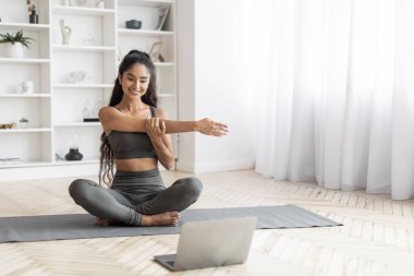 Atletik kıyafetlerle süslenmiş, kendini işine adamış genç bir Hintli kadın, evdeki açık dizüstü bilgisayarında sergilenen bir rehberi takip ederek aktif olarak antreman yapıyor. Fitness, yoga, pilates