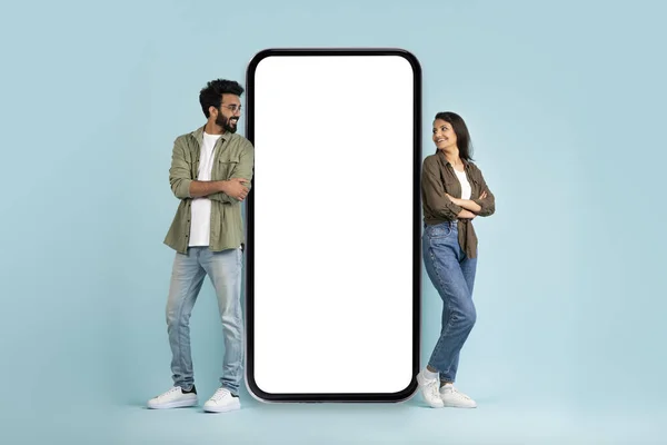 Dating mobile application. Happy millennial multiracial man and woman standing by big phone with white blank screen mockup for advertisement, looking at each other and smiling, copy space