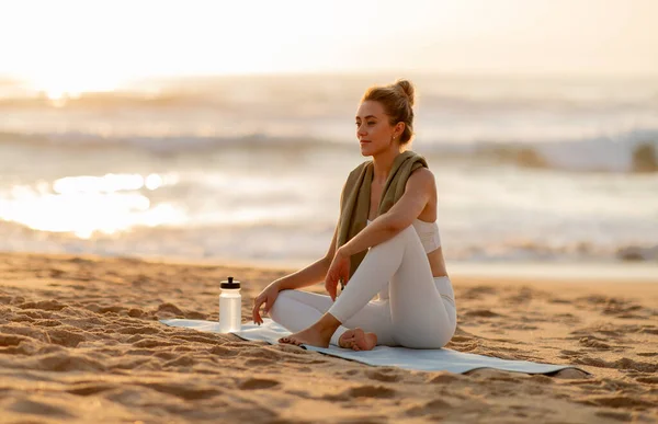Young woman relaxing after outdoor training, sitting on fitness mat on sandy sea beach and looking away, athletic lady resting after outside workout, free space