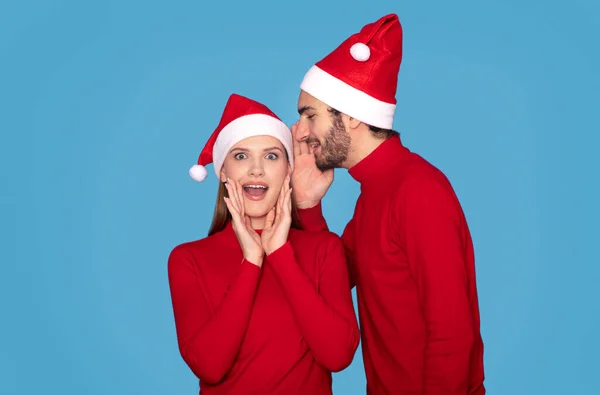 Christmas Offer. Young man whispering secret to his surprised wife, gossiping to her ear, couple wearing santa hats and standing over blue background, enjoying celebrating xmas holidays together