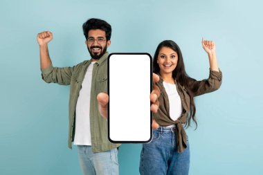 Online kuponlar, promosyon kodları, Cash Back. Duygusal heyecanlı genç Hintli çift beyaz ekranlı büyük bir telefon gösteriyor ve yumruklarını sıkıyor, mavi arka planda izole edilmiş, model, kopyalama alanı