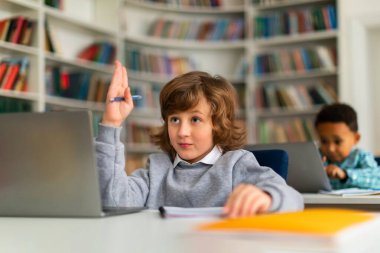 Çalışkan öğrenci, canlı bir ilkokul sınıfında dizüstü bilgisayarın önünde oturuyor, katılmak için elini kaldırıyor, ders sırasında katılma ve öğrenme hevesini sergiliyor.