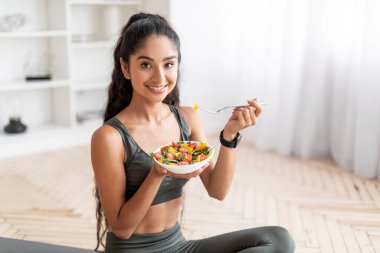 Spor kıyafetli neşeli, çekici genç Hintli kadın yoga minderinde oturuyor, antrenmandan sonra taze sebze salatası yiyor, ev içi, yakın çekim, fotokopi odası. Sağlıklı yaşam tarzı, diyet, spor