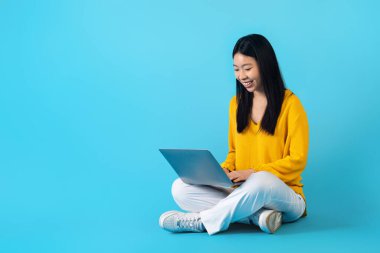 Gülümseyen, güzel, Asyalı bağımsız bir kadın, dizüstü bilgisayarda çalışıyor, mavi stüdyo arka planında yerde oturuyor. Çinli kadın çevrimiçi çalışıyor, bilgisayar kullanıyor, boşluğu kopyalıyor