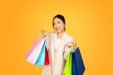 Alışveriş çılgınlığı yaşayan neşeli Asyalı kadın, kolları renkli çantalarla dolup taşıyor, hem alışverişin zevkini yansıtıyor hem de günümüz gençlerinin tüketici odaklı yaşam tarzını yansıtıyor.