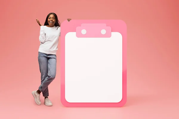 Shocked happy millennial black lady in casual with open mouth near big plan with free space, has fun, isolated on pink studio background. Day planning, surprise, motivation, success and win