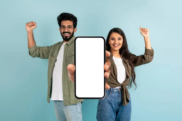 Online Coupons, Promo Codes, Cash Back. Emotional excited young indian couple showing big phone with white blank screen and clenching fists, isolated on blue background, mockup, copy space