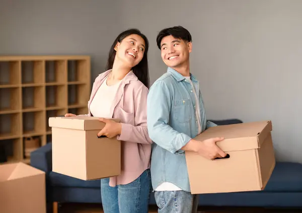 Real Estate Offer. Happy Asian Spouses Posing With Carton Boxes Holding Packed Belongings Moving in Their New Home, Standing Back to Back And Smiling To Each Other. Homeowners Concept