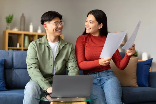 Easy Paperwork. Smiling Korean Spouses Using Laptop And Holding Bills Papers, Sitting On Couch In Cozy Living Room At Home. Young Couple Reading Documents And Letters. Good Financial News