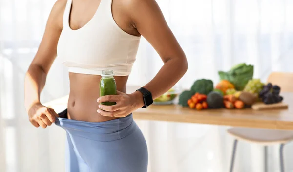 Young african american woman in sportswear, show weight loss result on pants, hold jar of fresh smoothie, juice on kitchen light interior with fresh vegetables. Diet at home, healthy food