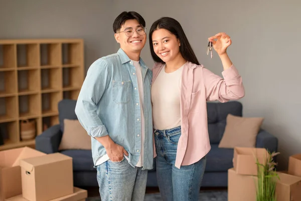 Joyful chinese couple proudly showcasing their new house keys, amid unpacked moving boxes, standing in their home. Husband hugging wife happy about real estate property purchase