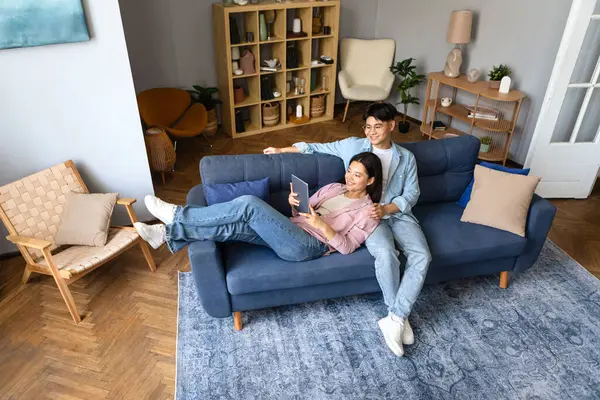 Asian man and woman browsing internet on their tablet computer in cozy modern living room, resting on sofa and having relaxed technology time on weekend, high angle. Website advertisement, gadgets