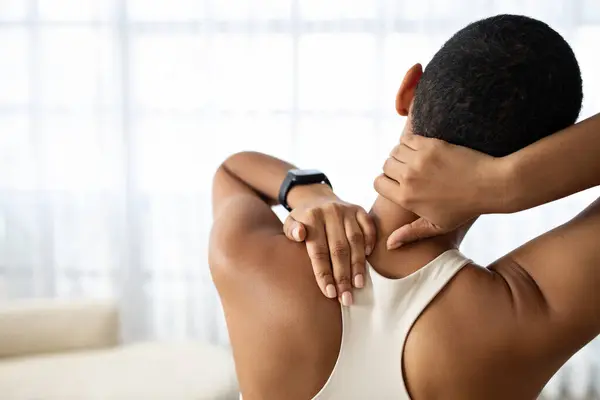 Young african american lady in sportswear massaging neck in living room interior. Sports at home, lady suffering from pain, injury after workout, muscle strain, inflammation, health problem