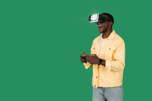 Young African American man wearing VR headset enjoy playing video game on green background. Black guy using VR goggles and joystick while experiencing the virtual reality, gaming, copy space