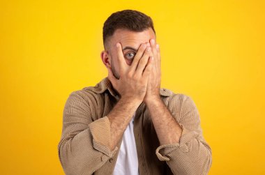 Bin yıllık Avrupalı bir adam, yüzünü ellerle kapatıyor, parmaklarından bakıyor, sarı stüdyo arka planında izole edilmiş, serbest alan. Sorunlar, stres, korku duyguları, korku filmi, yüz ifadesi