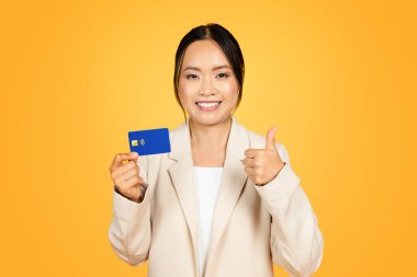 Takım elbiseli mutlu Asyalı kadın, elinde kredi kartı, baş parmak hareketi, tüketici karar verme, alışveriş onayı, tavsiye, turuncu arka planda izole edilmiş.