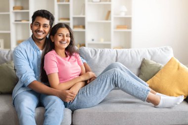 Keyifli Hintli çift, rahat bir evde, kanepede oturuyor, karısını kucaklıyor ve kameraya gülümsüyor, sıcak ve sevgi dolu, özgür bir yer.