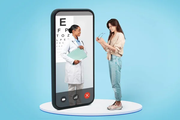 Smiling black lady doctor, specializing as ophthalmologist, expertly assisting patient in selecting new eyeglasses on big phone screen, isolated on blue background studio. Vision improvement app