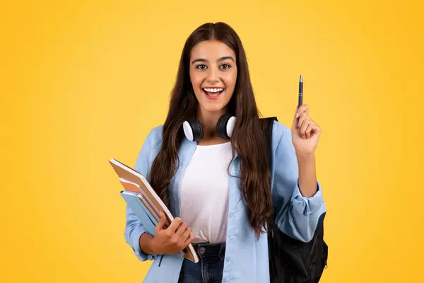 Cheerful european woman student, with books and headphones, rise pen up, got idea sign, isolated on yellow studio background. Education, good news emotions, create solution at school project