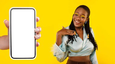 Gülümseyen siyah milenyum bayanı elini kendine doğrultuyor, akıllı telefonu boş ekranlı, sarı stüdyo arka planında izole edilmiş. Uygulama, web sitesi tavsiyesi, moda, güzellik bakımı