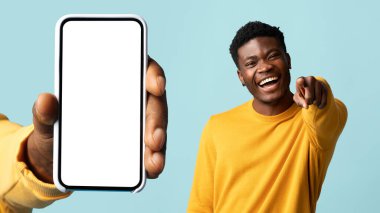 Gülümseyen Afro-Amerikan adam kamerayı işaret ediyor, telefonu boş ekran ile yakın el, mavi arka planda, stüdyoda, kolajda izole edilmiş. Eğlence, mizah uygulaması, web sitesi