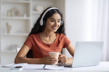 Kablosuz kulaklıklı, gülümseyen Hintli bir kadın masa başına oturmuş laptopunu yiyor ve kahve içiyor. Webinar izliyor, online sınıfa katılıyor, elinde kupa tutuyor, bedava fotokopi çekiyor.