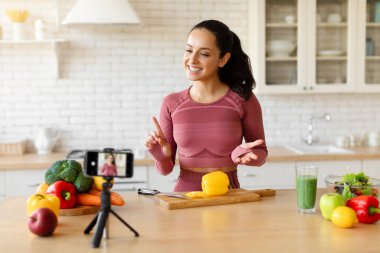 Beslenme blogu. Fitness blogcusu kadın mutfak içinde akıllı telefondan video pişiriyor, tripodla telefonla konuşuyor, sağlıklı beslenme tavsiyelerini ve yemek hazırlıklarını paylaşıyor.