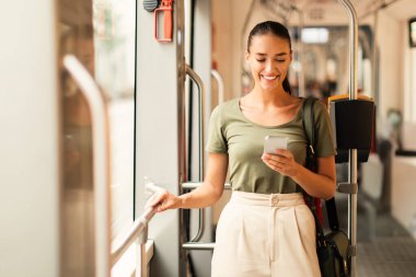 Gülümseyen genç bir kadın yolcu cep telefonunu kapalı bir tramvayda kullanıyor, internette geziniyor ve akıllı telefon uygulaması ile internet üzerinden tramvay bileti alıyor. Dijital teknoloji ve modern ulaşım