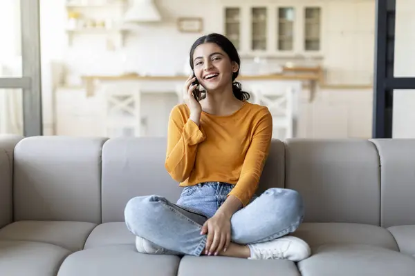 Mobile Call. Happy Young Indian Woman Talking On Cellphone At Home, Smiling Beautiful Eastern Female Enjoying Pleasant Phone Conversation While Sitting On Couch In Living Room, Copy Space
