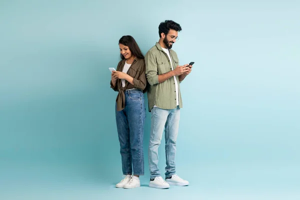 Dating mobile app concept. Stylish young multiethnic man and woman hipsters standing back to back, using smartphones on colorful studio background, chatting online, copy space, full length