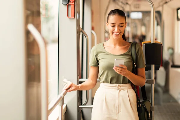 stock image Smiling young woman passenger using her mobile phone on a tram indoor, browsing web and booking streetcar ticket online via smartphone application. Digital technology and modern transportation