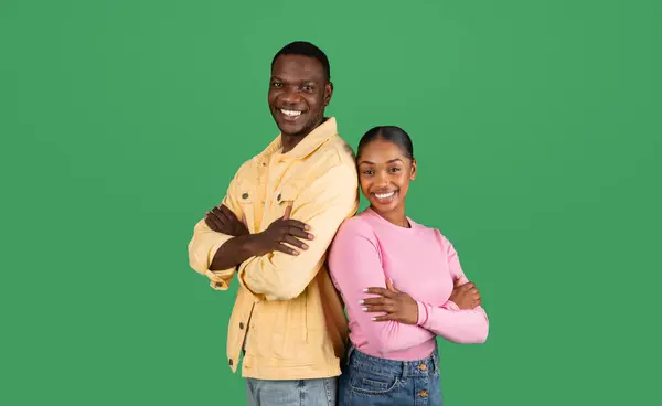 Cheerful smiling young black man and woman wearing casual outfits standing back to back, holding hands crossed on chest, isolated on green background. Teamwork, business, partnership