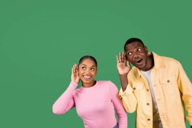 Meraklı genç siyah çift, yeşil stüdyo arka planında izole edilmiş bir şekilde gizlice dinliyor. Afrikalı Amerikalı erkek ve kadın, kulaklarının yanında el ele tutuşuyor, dedikoduları dinliyor, uzayı kopyalıyor.