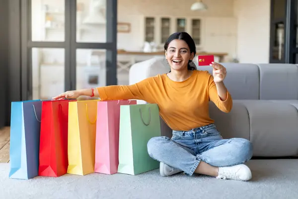 Happy Young Indian Woman Sitting Next To Bright Shopping Bags And Holding Credit Card, Cheerful Eastern Shopaholic Lady Enjoying Seasonal Sales And Discounts, Posing In Home Interior, Free Space