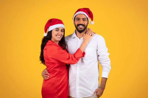 Glad young couple in Christmas hats, embracing and sharing festive moments, isolated on yellow background. Joy and togetherness during holiday season, love and celebration together
