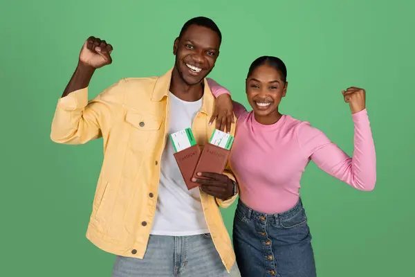 Excited happy young black people tourists cheerful man and woman showing tickets and passports, gesturing clenching fists on green background, going vacation. Tourism, journey, traveling